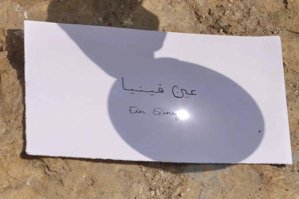 thesun still einqiniya sunshine through a magnifying glass on a paper, letting appear a text in arabic and english saying "Ein Qiniya"